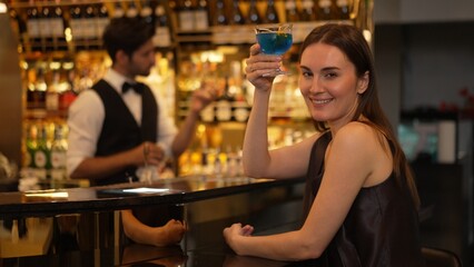 Sitting beautiful woman guest looking at camera to pose with drinking mixed vodka in blue hawaii cocktail at luxurious counter bar for Friday meeting on smart bartender background at club. Vinosity.