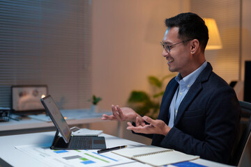 Asian businessman sitting at his desk in the office late at night, gesturing during a video call on his tablet