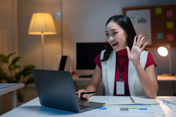 Asian businesswoman is working late and waving at someone during a video call on her laptop