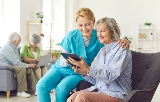 Caring kind smiling nurse, geriatric medicine caretaker watching photos or videos online on digital computer tablet providing senior woman help, support, aged old granny medical visit, female doctor 
