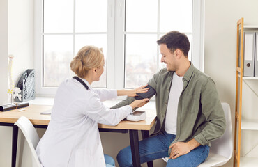 Young blonde female doctor therapist measuring blood pressure of cheerful male patient sitting in...