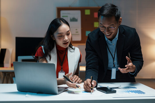 Two young professionals discussing a project, looking at charts and using a laptop, working late in the office