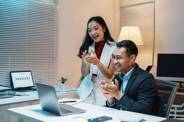 Two business people clapping their hands while looking at a laptop screen during a late night work session