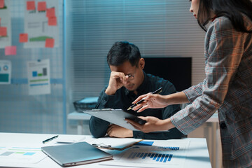 Businesswoman pointing at financial document while helping stressed businessman working late in office