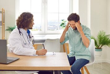 Portrait of a sick stressed young man sitting at the doctor's office suffering from headache or having migraine. Male patient during medical examination in clinic. Health care and medicine concept.