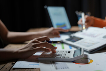 Business people analyzing financial documents or charts calculating company revenue using calculator and laptop at night in office