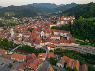 Panoramic view of historic Skofja Loka town in the valley with medieval castle on the hill. Aerial view above bus station and bridges over Sora river at sunset. Beautiful hilly green landscape.