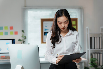 Asian businesswoman is standing in an office setting, writing on a clipboard while holding it in her hands