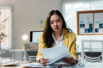 Asian businesswoman in a yellow blazer is reviewing paperwork at her desk in a modern office