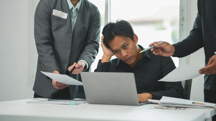 Young businessman is looking stressed while working on a laptop as his colleagues stand around him pointing at documents ©  NCST Studio