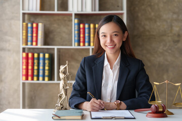 Lawyer is smiling while working at her desk, with a statue of lady justice and a gavel in the...