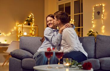 Young man congratulating pretty smiling happy woman on holiday with red present gift box hugging and kissing her sitting on sofa at home. Couple in love, tenderness and Valentines day concept.