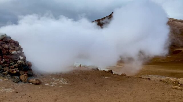 White steam billows from a geothermal vent, rising through the rugged landscape of Hverir near the Kafla vulcanic area of Iceland, adorned with rocky formations and towering mountains