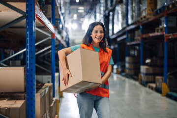 Female warehouse worker carrying cardboard box and smiling at logistics center