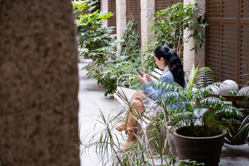 A beautiful adult woman scrolls on her phone sitting on a bench on a stone patio with classic...