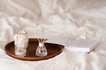 Liquid home fragrance in glass bottle with bamboo sticks with scented candle on tray in bed over white close up. Cozy atmosphere. Aromatherapy.