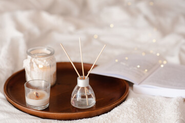 Liquid home fragrance in glass bottle with bamboo sticks with scented candle on tray in bed over white close up. Cozy atmosphere. Aromatherapy.