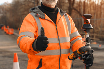 A skilled roadworker gives a thumbs up while using surveying equipment on a construction site in autumn