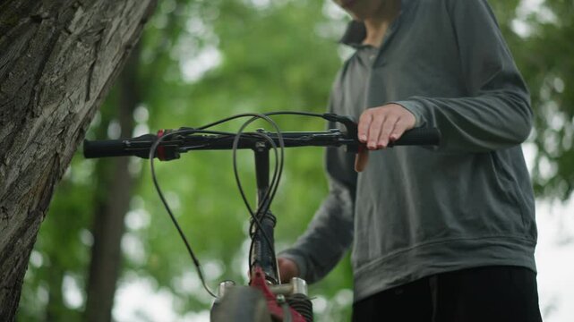 Boy wearing glasses firmly grips the handlebar of parked bicycle near tree in grassy field, he is checking the brake functionality while standing, his face is blurred in the background