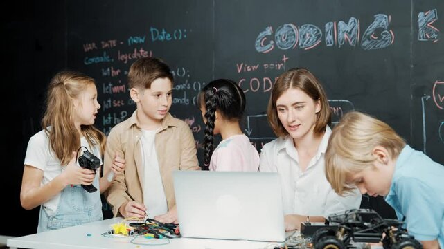 Caucasian teacher using laptop and explain about engineering system while diverse student holding electronic equipment in front of blackboard with prompt or code written in STEM class. Erudition.
