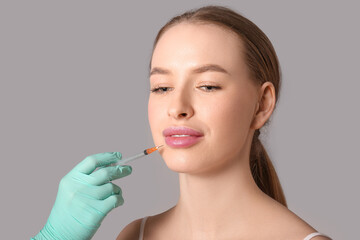 Young woman receiving filler injection in lips on grey background