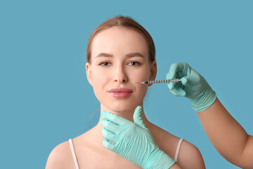 Beautiful young woman receiving filler injection in face on blue background