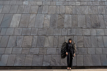 A mysterious figure with wings stands against a modern stone wall in an urban setting at dusk