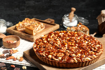 Wooden board of sweet tart with different nuts and caramel on black background
