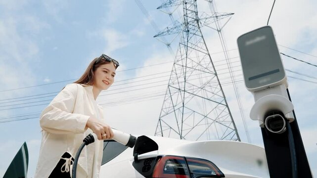 Bottom view slow motion woman recharge EV car battery at charging station connect to power grid tower electrical industrial facility as electricicty industry for eco friendly vehicle. Expedient