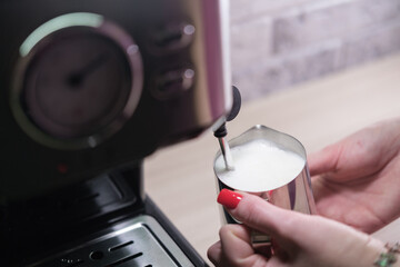 Girl holds pitcher with milk and heats it with coffee machine. Milk for making cappuccino. Close-up. Making coffee at home