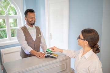 A smiling hotel clerk assists a guest during checkout in a well-lit reception area. The guest is...