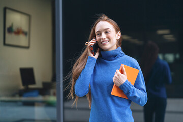 A young woman smiling while talking on the phone and holding a notebook outside a modern office...