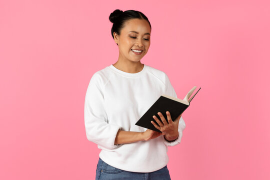 A young Asian woman stands joyfully reading a book, her expression vibrant and engaged. The backdrop is a solid bright pink, enhancing her cheerful demeanor.