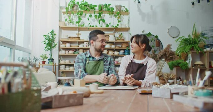 Front camera view of Caucasian couple working together as potters in bright room. Helping each other on busy day. Wedging clay. Kneading material required for creation of pots or cups.