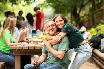 A joyful family gathering is happening in a park. A man is hugging a young woman, both smiling, surrounded by loved ones enjoying food and drinks on a sunny day.