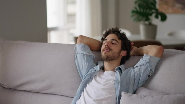 Happy relaxed young man rest lounge lean on couch enjoy peaceful mood, healthy lazy calm guy dreaming breathing fresh air sit on comfortable sofa in living room on stress free cozy day at home
