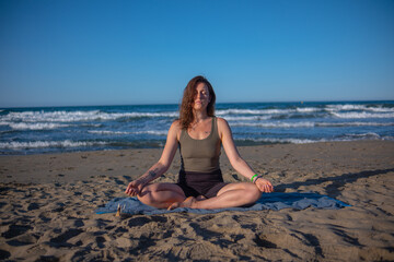 Girl in sportswear doing yoga on sea sand beach. Morning yoga on seashore under clear blue sky. Sporty young woman maintains healthy lifestyle