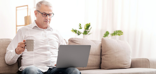 Serious Mature Man Holding Laptop Computer And Coffee Mug, Sitting On Sofa At Home, Empty Space
