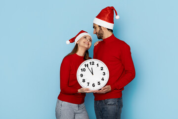 A joyful couple in matching red sweaters holds a large clock, waiting for midnight. Both wear Santa...