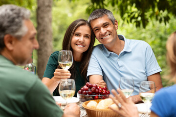 A couple smiles while toasting with glasses of wine at a festive outdoor gathering. Friends relax around a table filled with fruit, bread, and laughter under leafy trees.