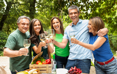 A cheerful gathering of five friends toasting with glasses of wine at a picnic table filled with fresh produce, surrounded by lush trees on a sunny day.