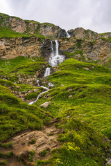 Photo of waterfall in mountains. Rapid streams of water cascade down steep cliffs surrounded by greenery. Majestic Alpine peaks. Grossglockner High Alpine Road. Austria