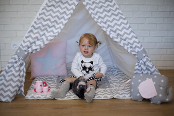 Adorable beautiful happy smiling ginger-haired toddler little girl is playing with her toys sitting in a teepee wigwam © Tina