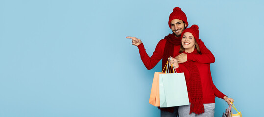 A cheerful couple stands together, both wearing red hats and sweaters, while holding colorful shopping bags. Their joyful expressions capture the spirit of a fun shopping day, copy space
