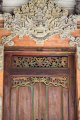 An intricately carved wooden door with ornate decorative details on top, featuring a traditional Balinese design. The carvings above the door depict a Barong face and floral motifs.