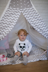 Adorable beautiful happy smiling ginger-haired toddler little girl is playing with her toys sitting in a teepee wigwam © Tina