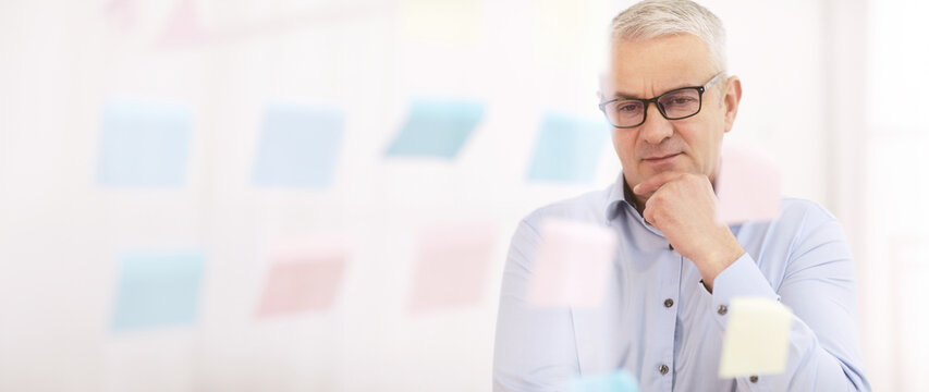 Brainstorming. Thoughtful mature businessman in specs looking at sticky notes on glass wall, touching chin, empty space
