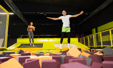 Cheerful young man enjoying soft landing in foam pit at trampoline park, with friends jumping around