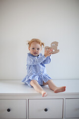 Aesthetic minimalistic portrait of Adorable beautiful smiling happy ginger-haired toddler baby girl sits on a white chest of drawers and plays with a toy horse
