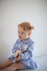 Aesthetic minimalistic portrait of Adorable beautiful smiling happy ginger-haired toddler baby girl sits on a white chest of drawers and plays with a toy horse